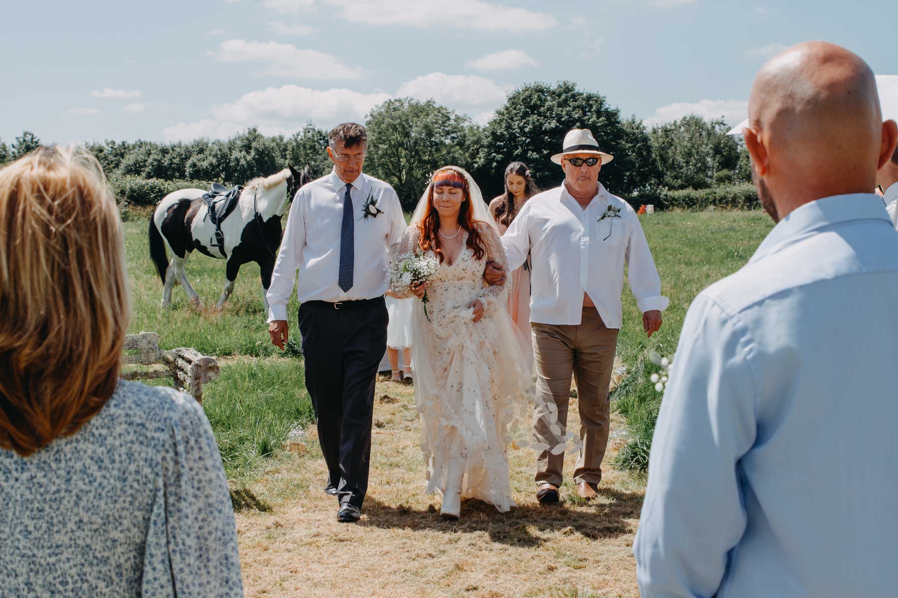 The bride walks down to the handfasting ceremony with her father and stepfather. Handfasting ceremony photography reportage