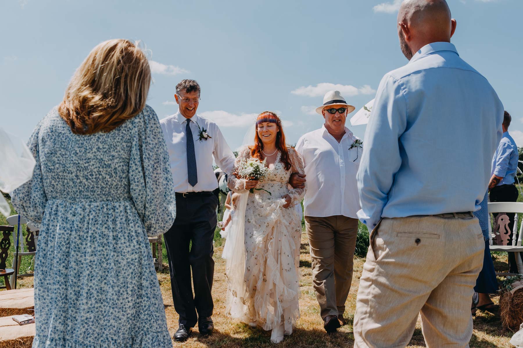 The bride with her father and stepfather approaches the groom and their celebrant of the handfasting ceremony. Handfasting ceremony photography reportage