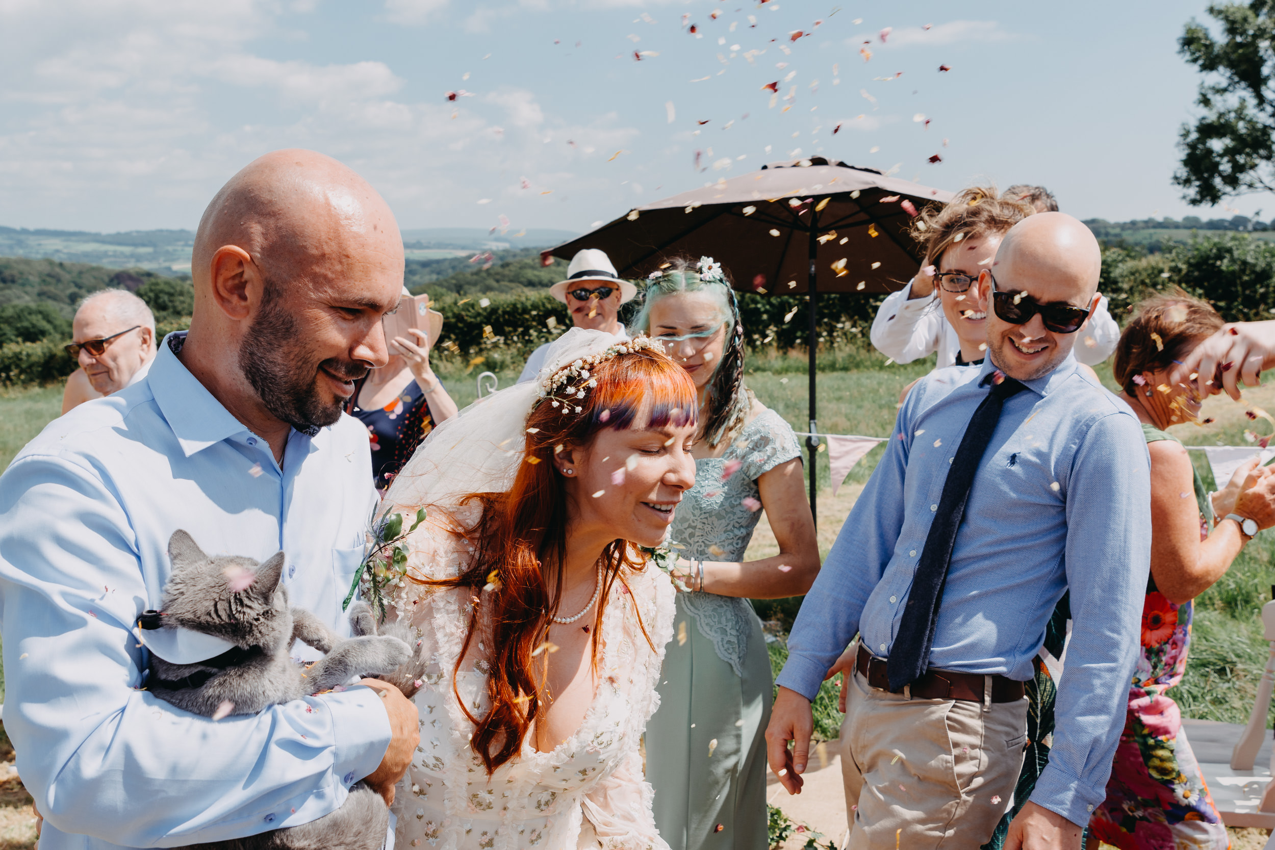 Bride and groom surrounded by joyful guests throwing confetti during a sunny outdoor farm wedding, captured by a candid wedding photographer.
