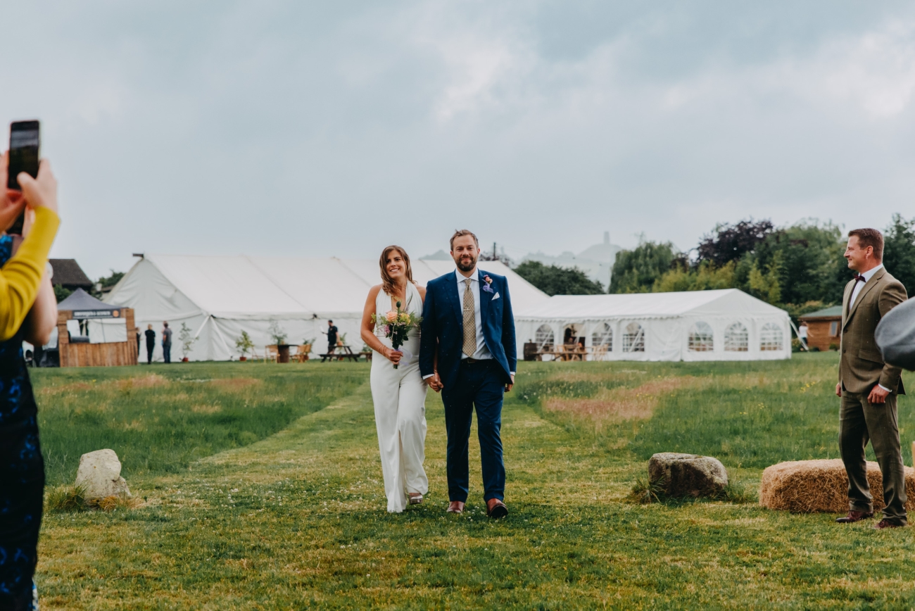 Bride and groom walking hand-in-hand down a grassy path towards the festival-style wedding setup at Splotts Moor, Glastonbury.