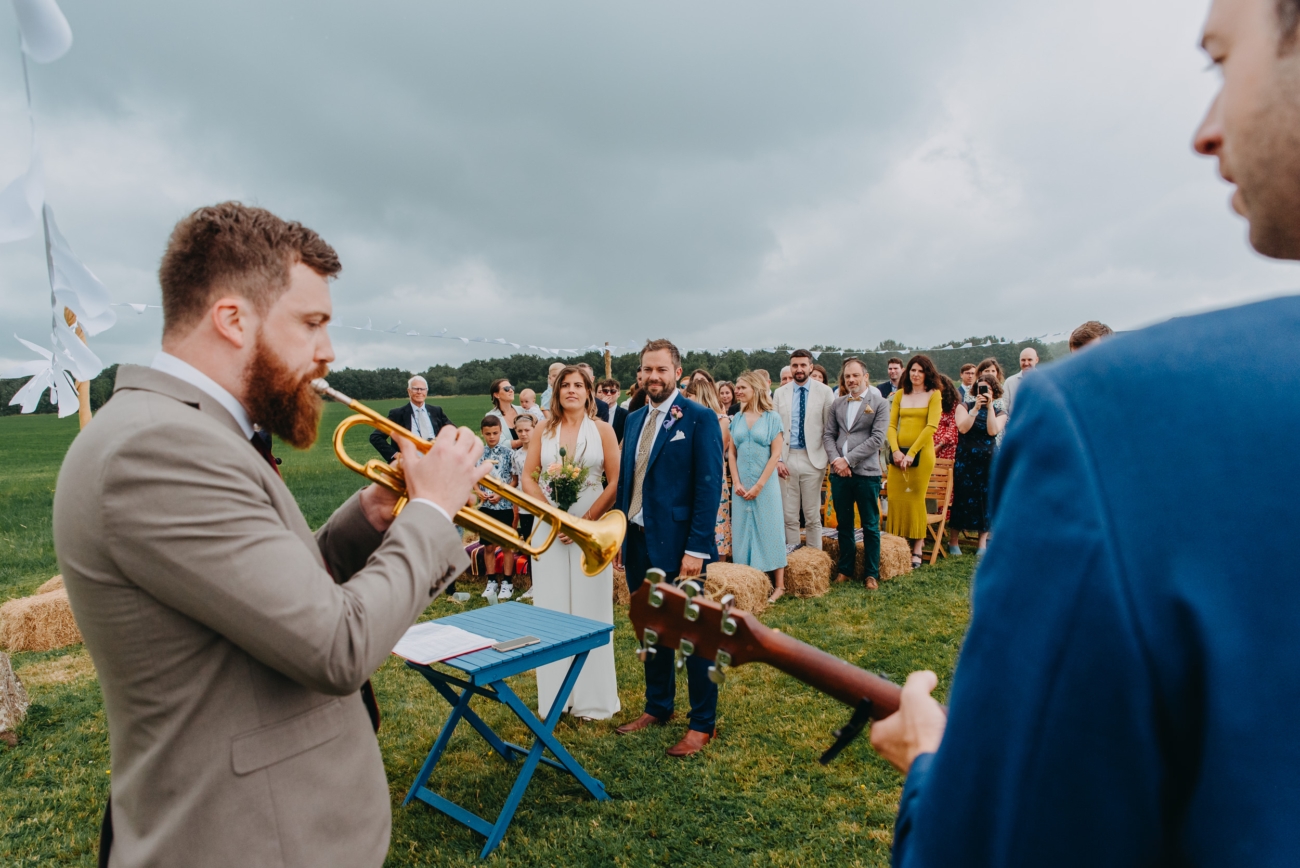Trumpet player performs as the bride and groom stand together during an outdoor festival-style wedding ceremony at Splotts Moor, Glastonbury, with guests watching in the background.