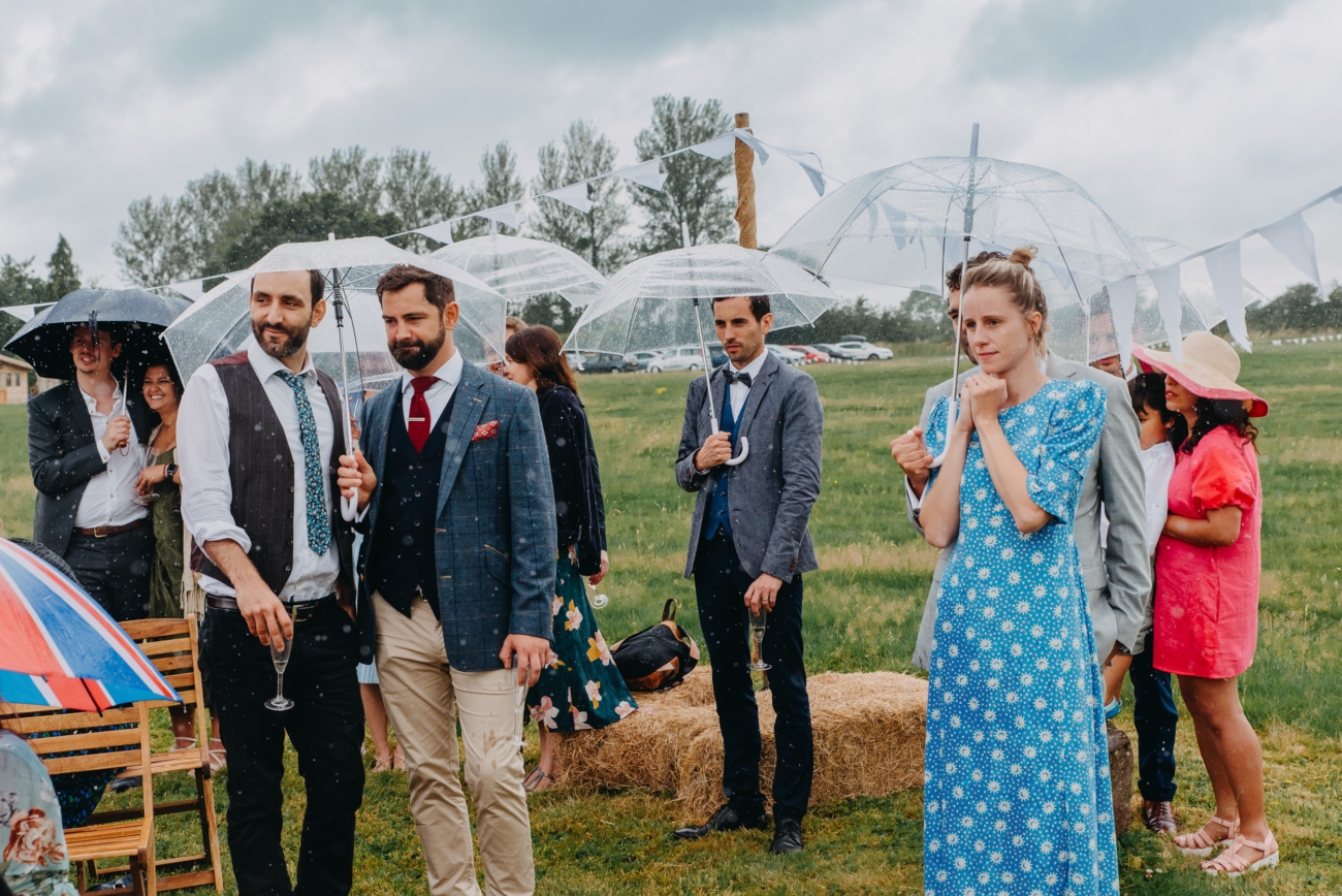 Guests stand under clear umbrellas, braving the rain with style and poise during the outdoor wedding ceremony, creating a quintessentially British festival wedding scene.