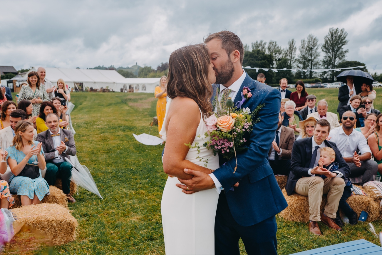 The bride and groom share a joyful first kiss after exchanging vows, with guests clapping and smiling in the background.