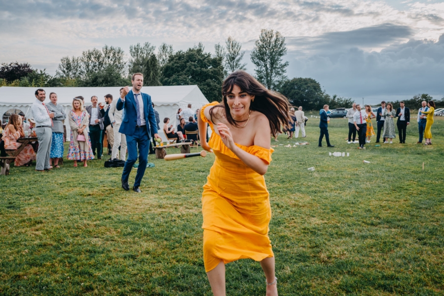 Wedding guest in a vibrant yellow dress enthusiastically participates in a game during an outdoor festival-themed wedding, captured by a candid wedding photographer.