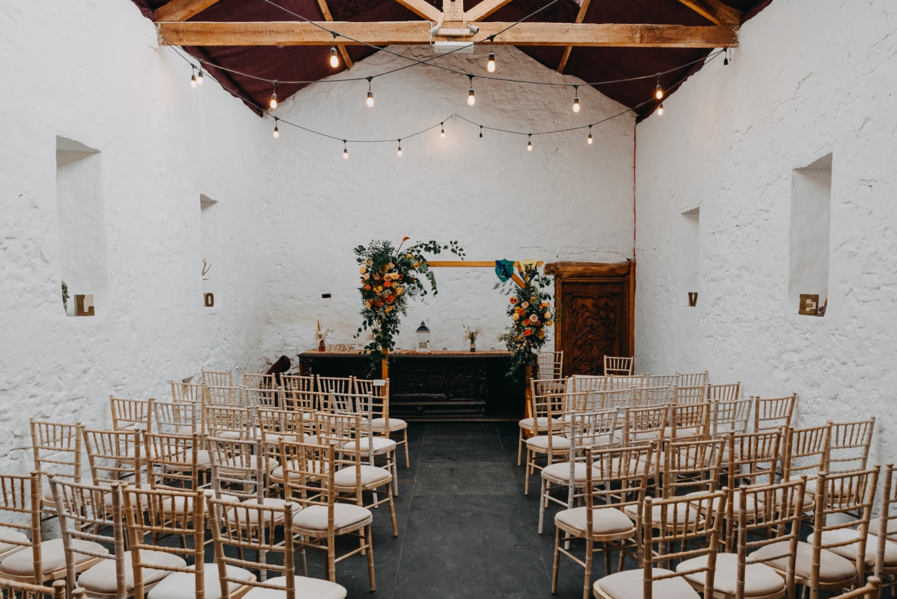 Elegant indoor wedding ceremony setup at Coed Weddings venue near Cowbridge, Wales, featuring rustic wooden beams, simple chairs, and vibrant floral arrangements.