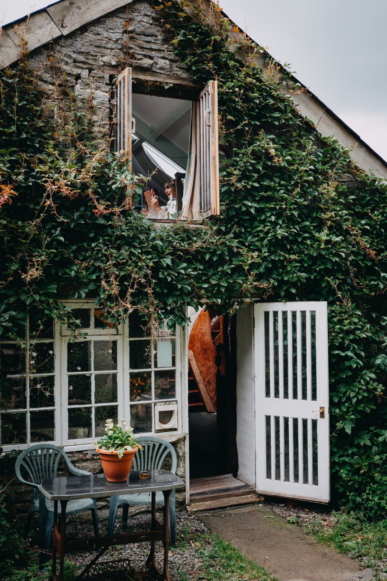 A bride getting ready, captured through an open window of an ivy-covered rustic cottage at Coed Weddings in Cowbridge, Wales. Below, a quaint outdoor seating area with potted plants adds to the serene, natural charm of the venue.