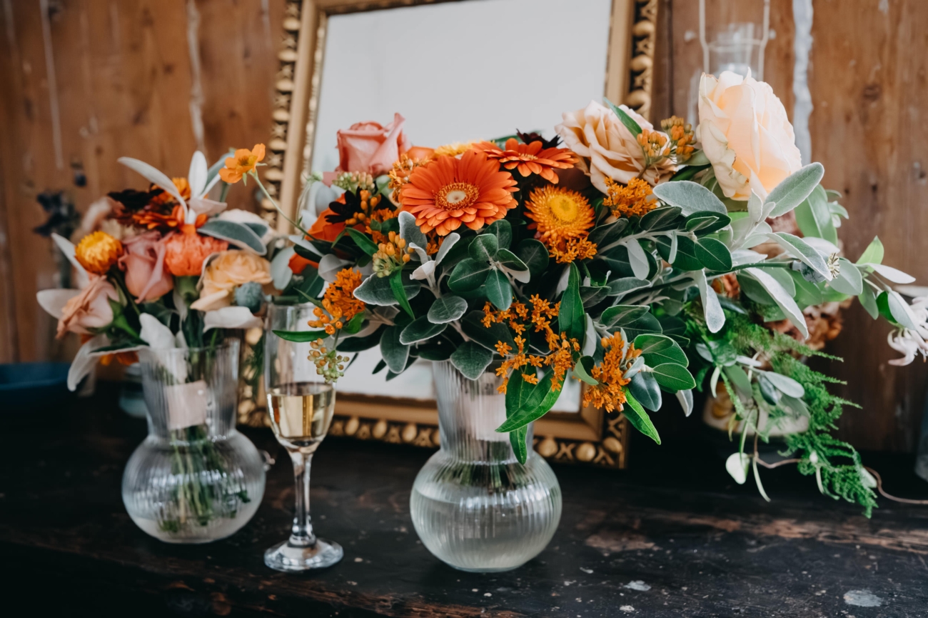 A close-up of vibrant floral arrangements featuring orange gerbera daisies, roses, and greenery in glass vases, with a champagne glass placed beside them, set against a rustic mirror at Coed Weddings, Cowbridge.