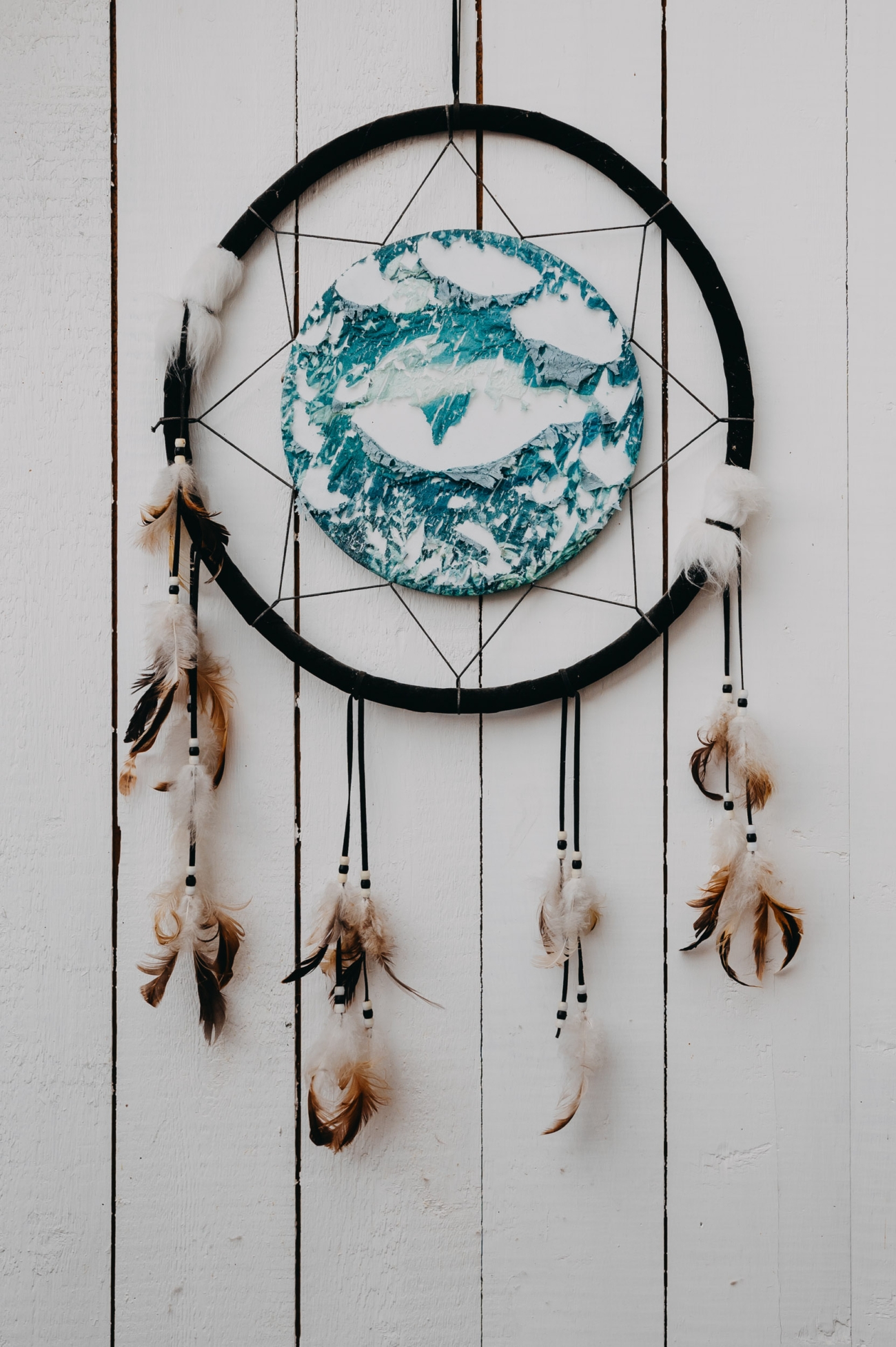 A handcrafted dreamcatcher with a circular frame, feathers, and a central design of blue and white abstract patterns, hanging against a rustic white wooden wall at Coed Weddings, Cowbridge.