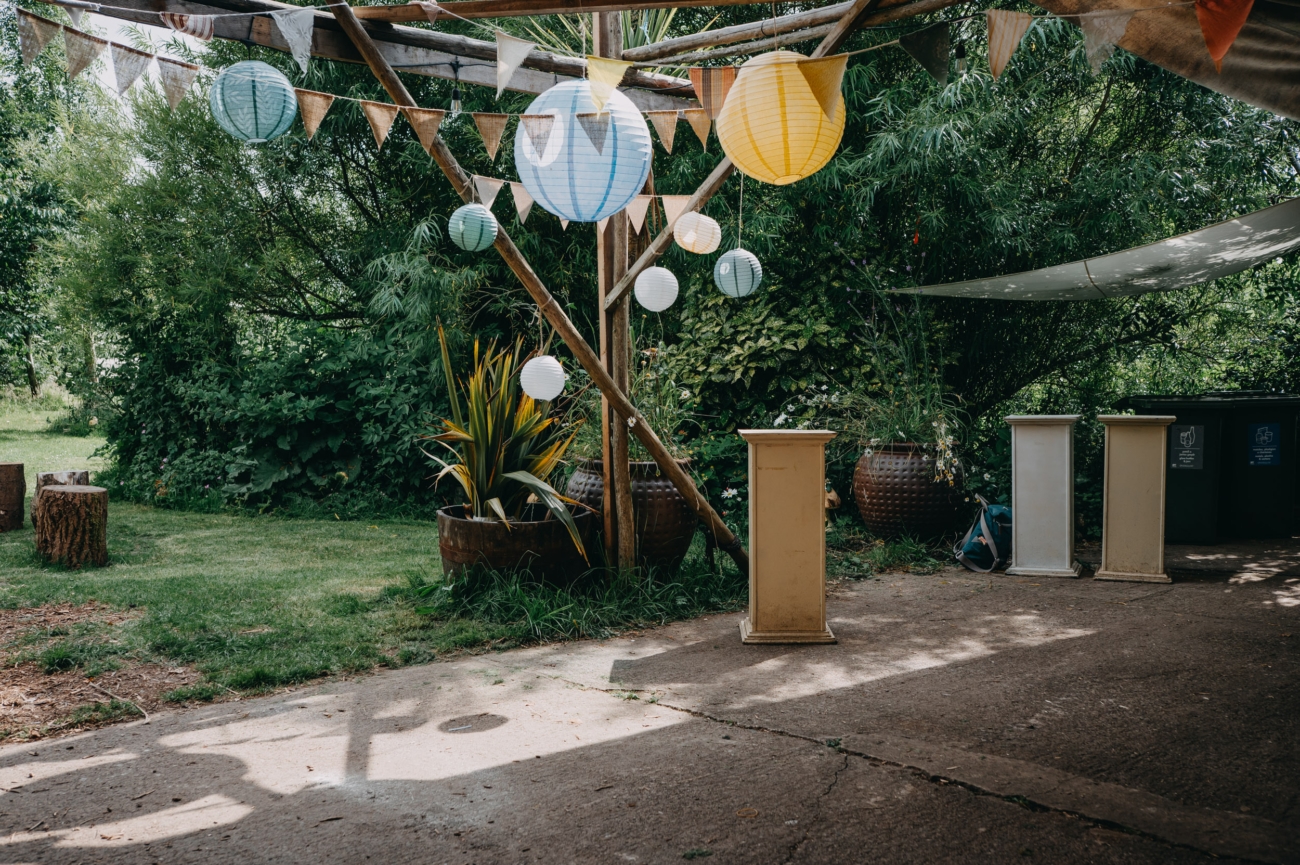 Outdoor wedding setup at Coed Weddings, Cowbridge, featuring colourful lanterns and bunting strung up on a wooden frame, with lush greenery in the background and rustic wooden stump seating in a relaxed, natural environment.