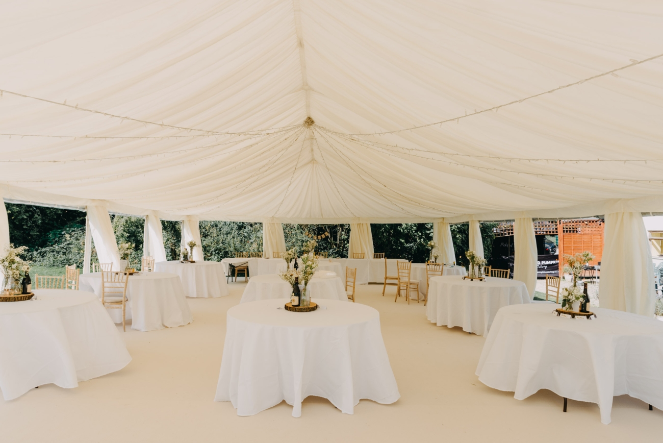 A beautifully decorated white marquee at Glansevin Mansion, Llangadog, set up for a wedding reception, with round tables draped in white cloths, minimalist floral centrepieces, and elegant wooden chairs.