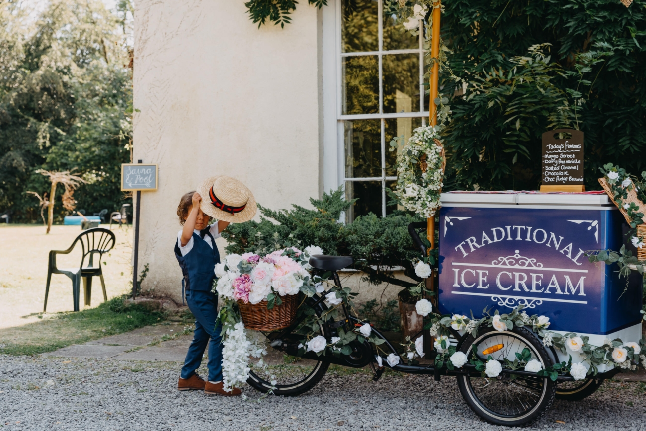 A young boy at Glansevin Mansion wedding venue, adjusting a straw hat while standing beside a charmingly decorated traditional ice cream bike adorned with flowers.