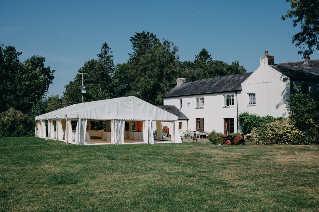 A white marquee tent set up on the green lawn beside the historic Glansevin Mansion in Llangadog, Wales, under a bright blue sky.