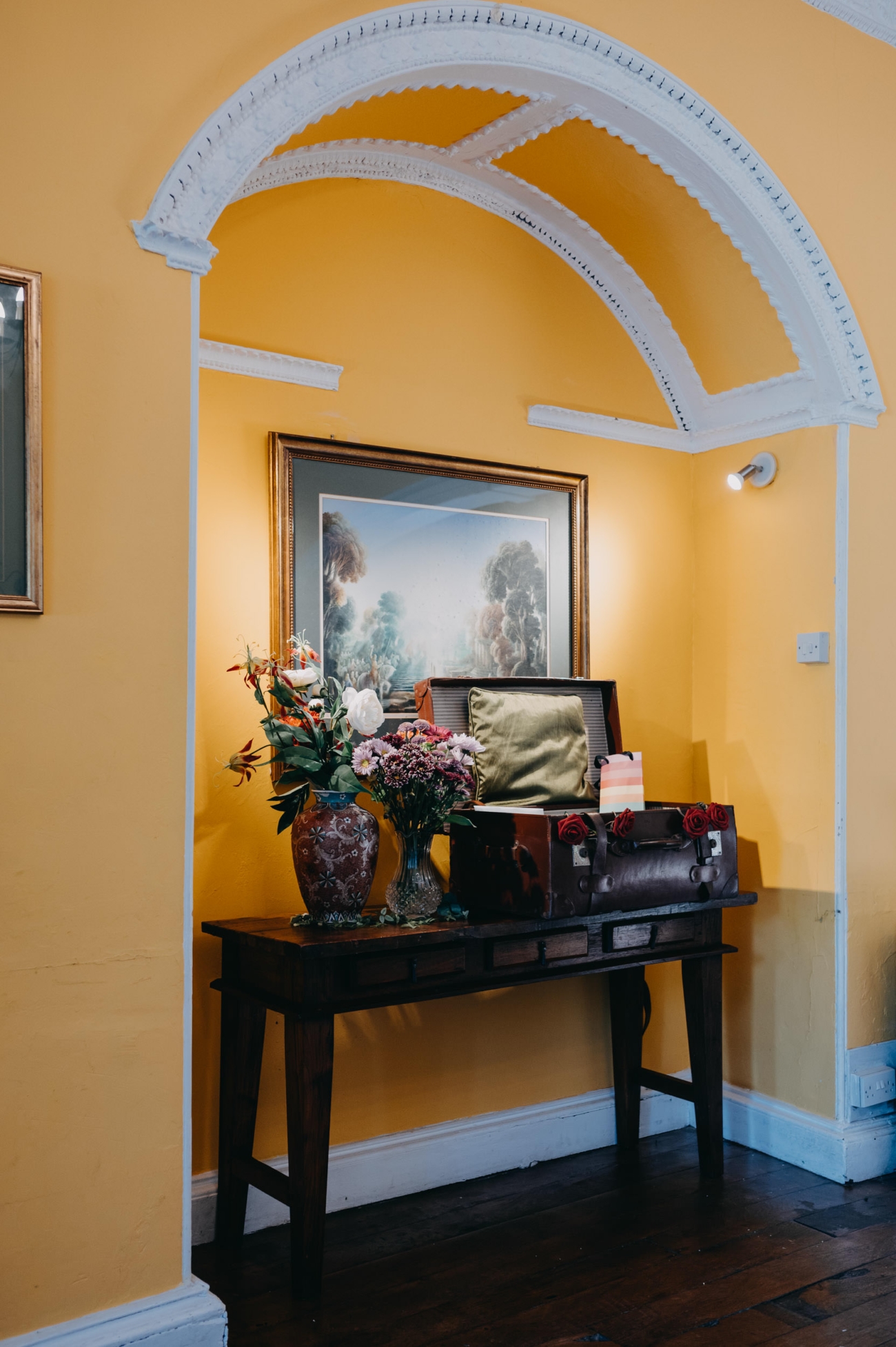 An elegantly arranged corner inside Glansevin Mansion, Llangadog, featuring a vintage wooden table adorned with a vase of colourful flowers, a decorative suitcase, and an ornate framed painting on a yellow wall under a white archway.