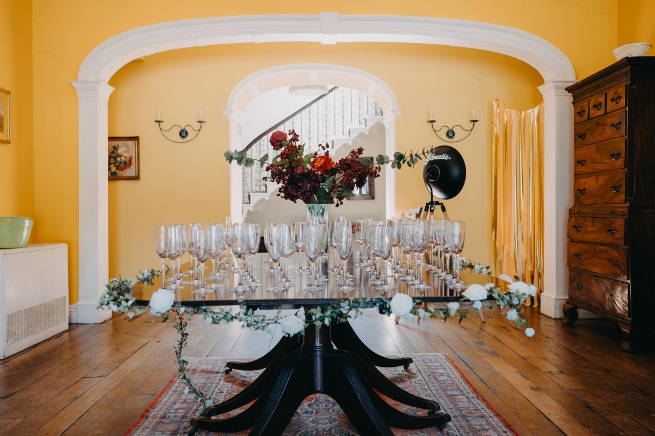 A beautifully set table at Glansevin Mansion, Llangadog, adorned with numerous empty wine glasses and a floral arrangement, placed beneath an archway. The yellow walls, elegant staircase in the background, and vintage wooden furniture add to the room’s sophisticated atmosphere.
