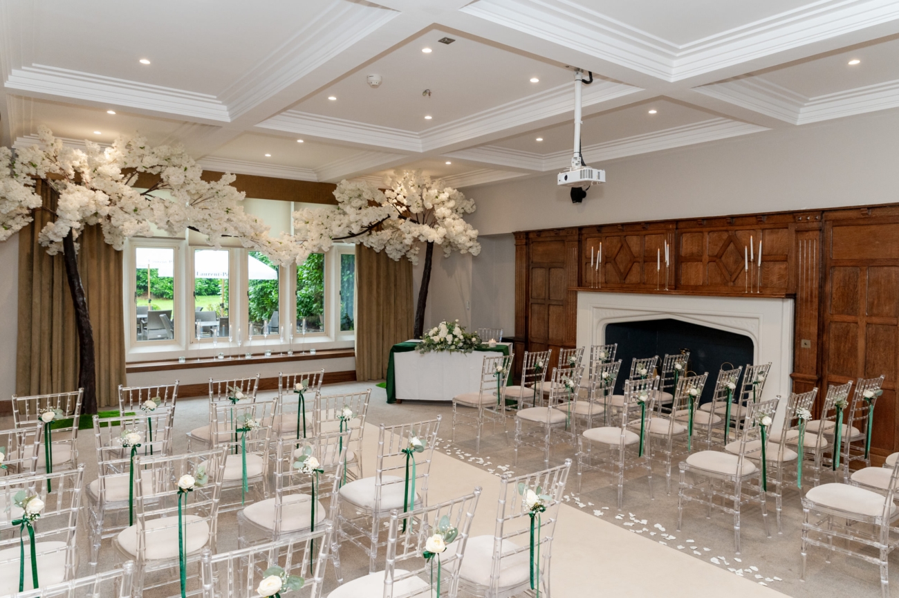 A beautifully decorated wedding ceremony room at Lanelay Hall. The space features transparent chairs adorned with white roses and green ribbons, while large faux cherry blossom trees frame the front of the room. The wood-panelled walls and fireplace add a touch of classic elegance, complementing the bright and airy atmosphere.