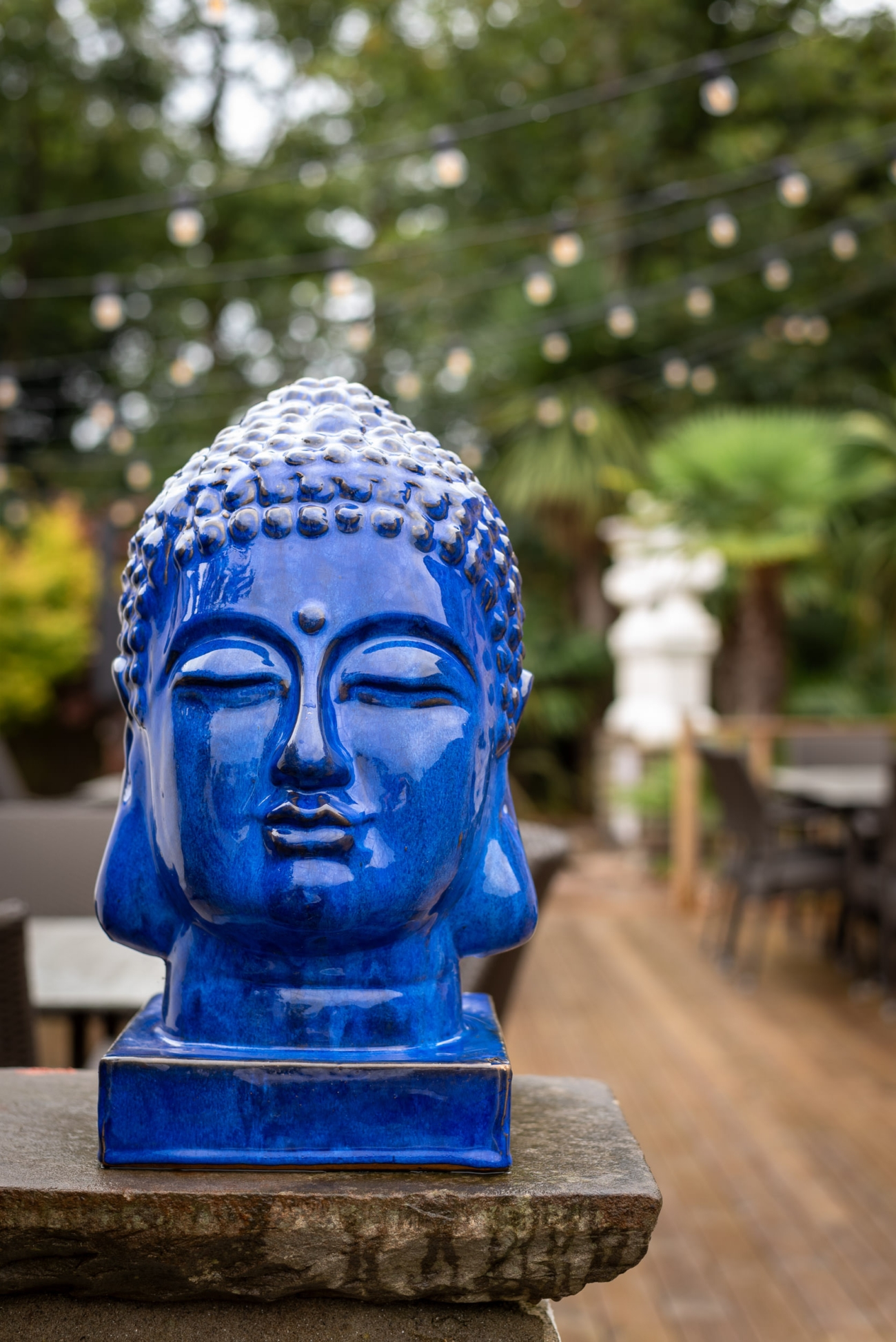 A striking blue Buddha statue on display at Lanelay Hall's outdoor area. The serene expression on the statue's face contrasts with the warm wooden decking and the soft glow of string lights hanging above. Lush greenery surrounds the space, adding to the tranquil and peaceful atmosphere.