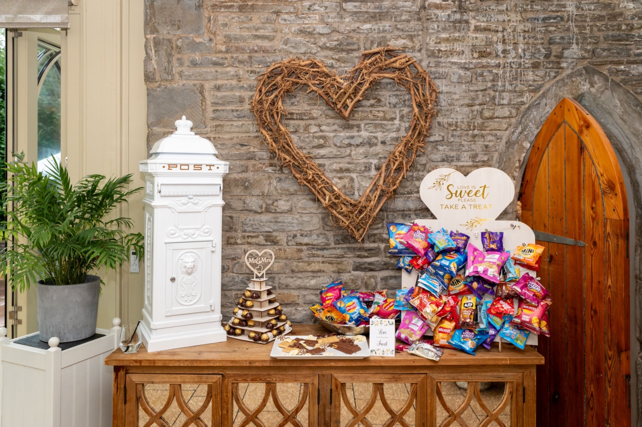 Wedding reception table at Lanelay Hall in Pontyclun, decorated with a white post box, a sweet treat station with snacks, and a rustic heart-shaped wreath on the stone wall.