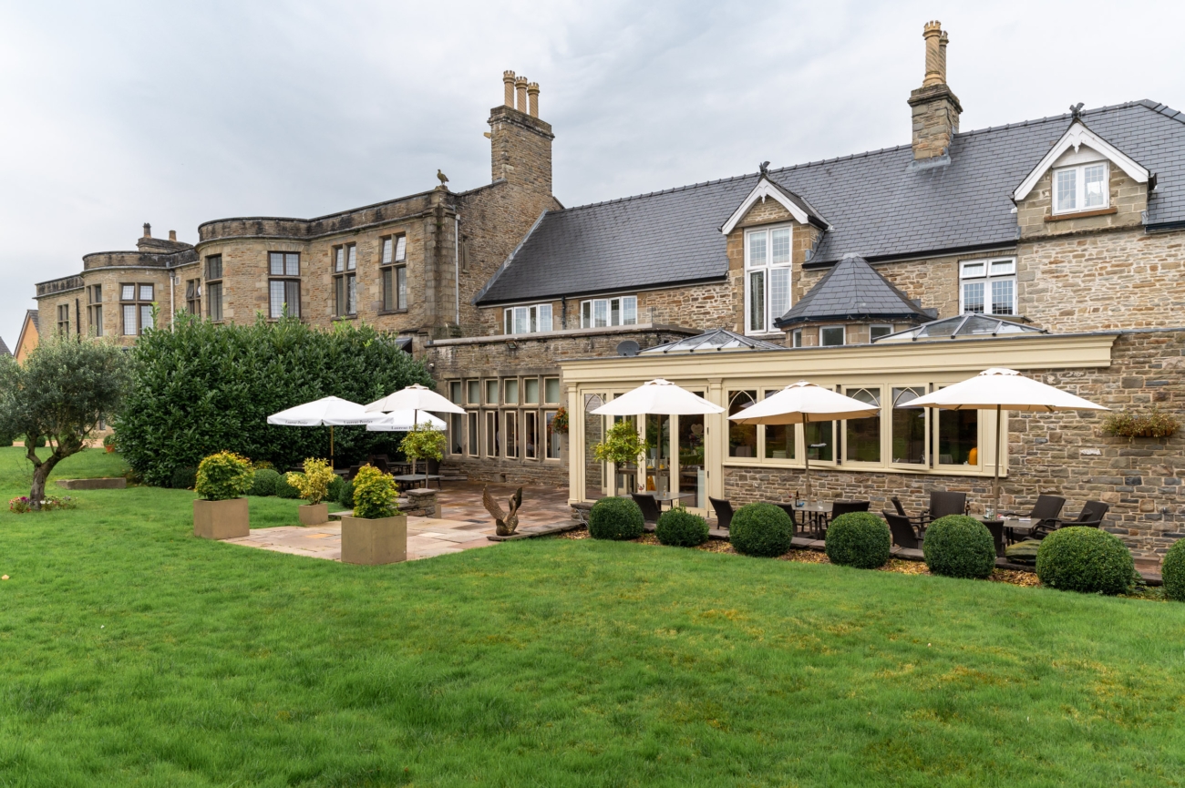 Elegant stone exterior of Lanelay Hall in Pontyclun, featuring a lush green lawn, outdoor seating with white parasols, and a mix of modern and historic architectural styles, perfect for outdoor wedding receptions.
