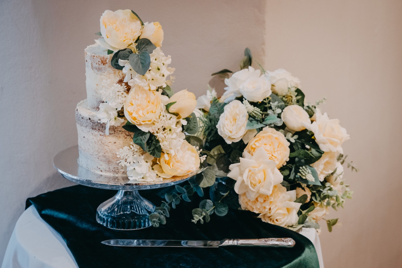 Elegant two-tiered semi-naked wedding cake adorned with soft yellow and white flowers, displayed on a crystal cake stand with a matching floral arrangement, ready for the cake cutting ceremony at Lanelay Hall in Pontyclun.
