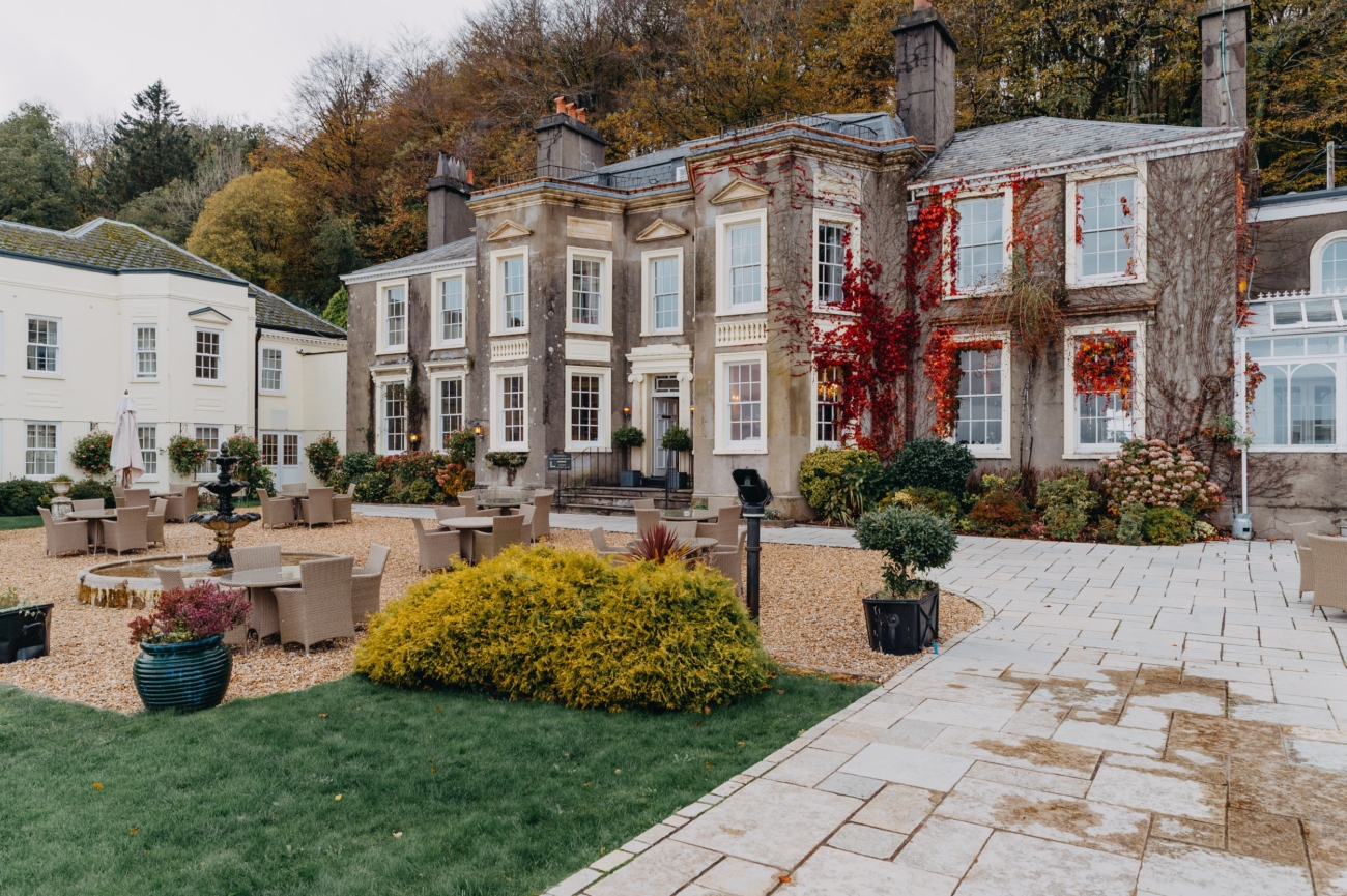 Stately exterior of the New House Country Hotel in Thornhill, Cardiff, surrounded by vibrant autumnal foliage and an elegant outdoor seating area with a central water fountain.