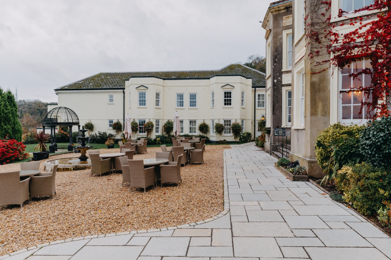 Outdoor seating area at New House Country Hotel in Thornhill, Cardiff, featuring wicker chairs and tables, a water fountain, and a garden pavilion in the background, with a backdrop of the hotel’s light cream façade.