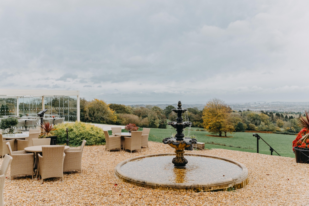Panoramic countryside view from the terrace of New House Country Hotel in Cardiff, featuring a central fountain, wicker seating, and a glass conservatory, with expansive green fields and the city of Cardiff visible in the distance.