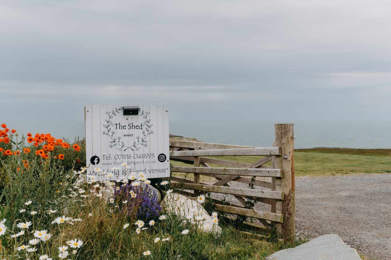Entrance sign for The Shed at Mwnt, a rustic wedding venue in Wales, surrounded by wildflowers and offering stunning sea views from a secluded location.