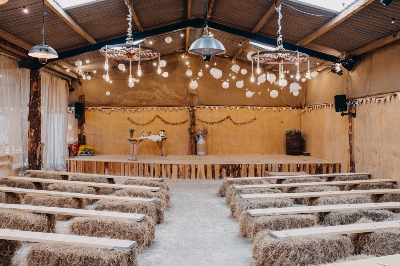 Rustic indoor setup at The Shed, Mwnt wedding venue, featuring hay bale seating, soft lighting, and a simple wooden stage for a charming countryside wedding.