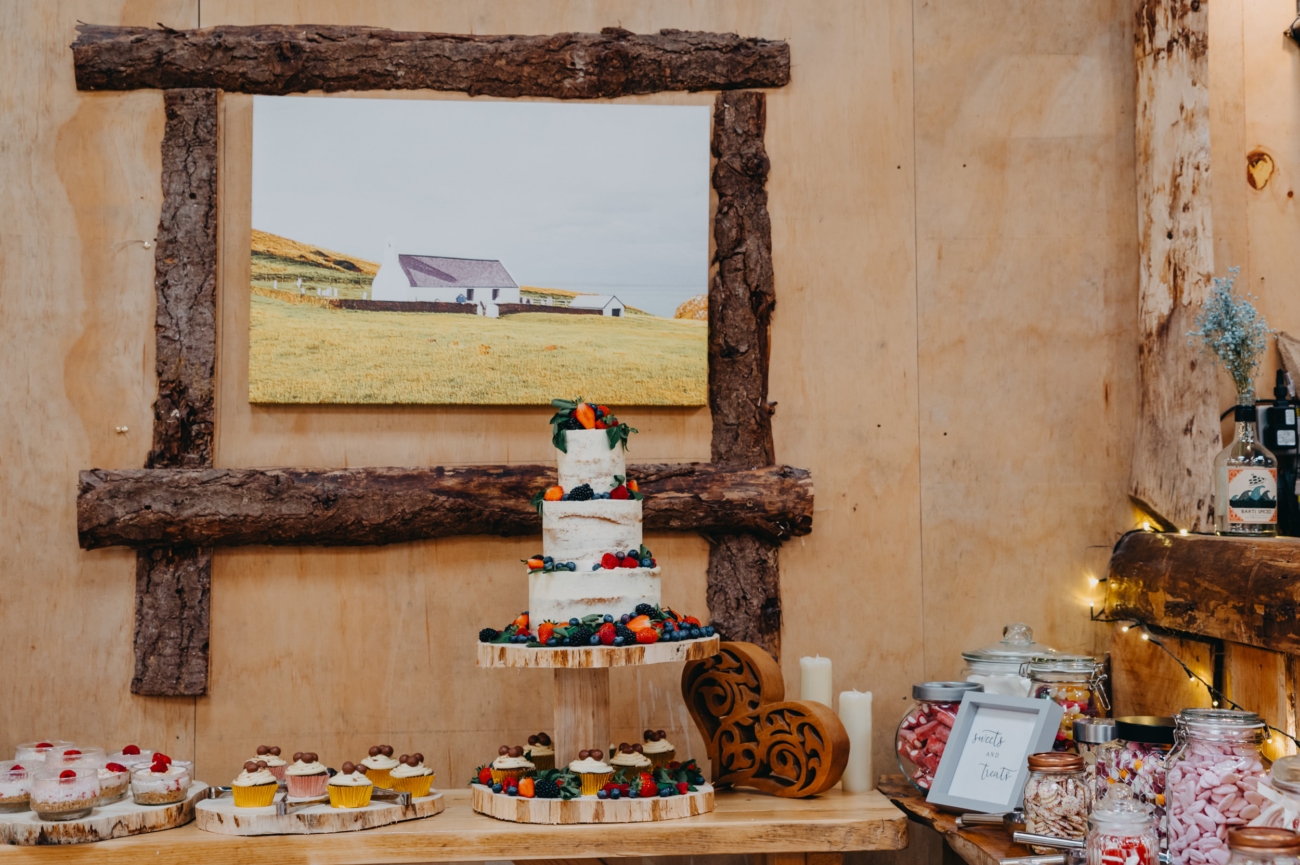 A rustic wedding cake display at The Shed, Mwnt, featuring a two-tiered naked cake with fresh fruit and a wooden backdrop, alongside cupcakes and a sweet treats station.