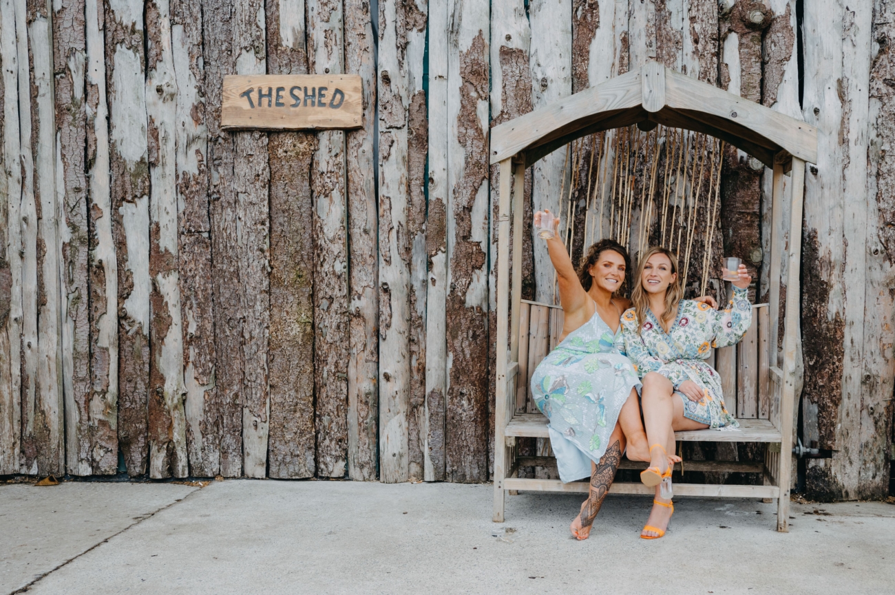 Two guests sitting on a wooden swing at The Shed, Mwnt, celebrating with drinks in hand in front of the rustic barn exterior.