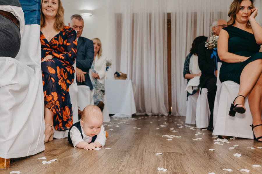 A baby crawling on the floor during a wedding ceremony, surrounded by flower petals, candidly captured among seated guests.