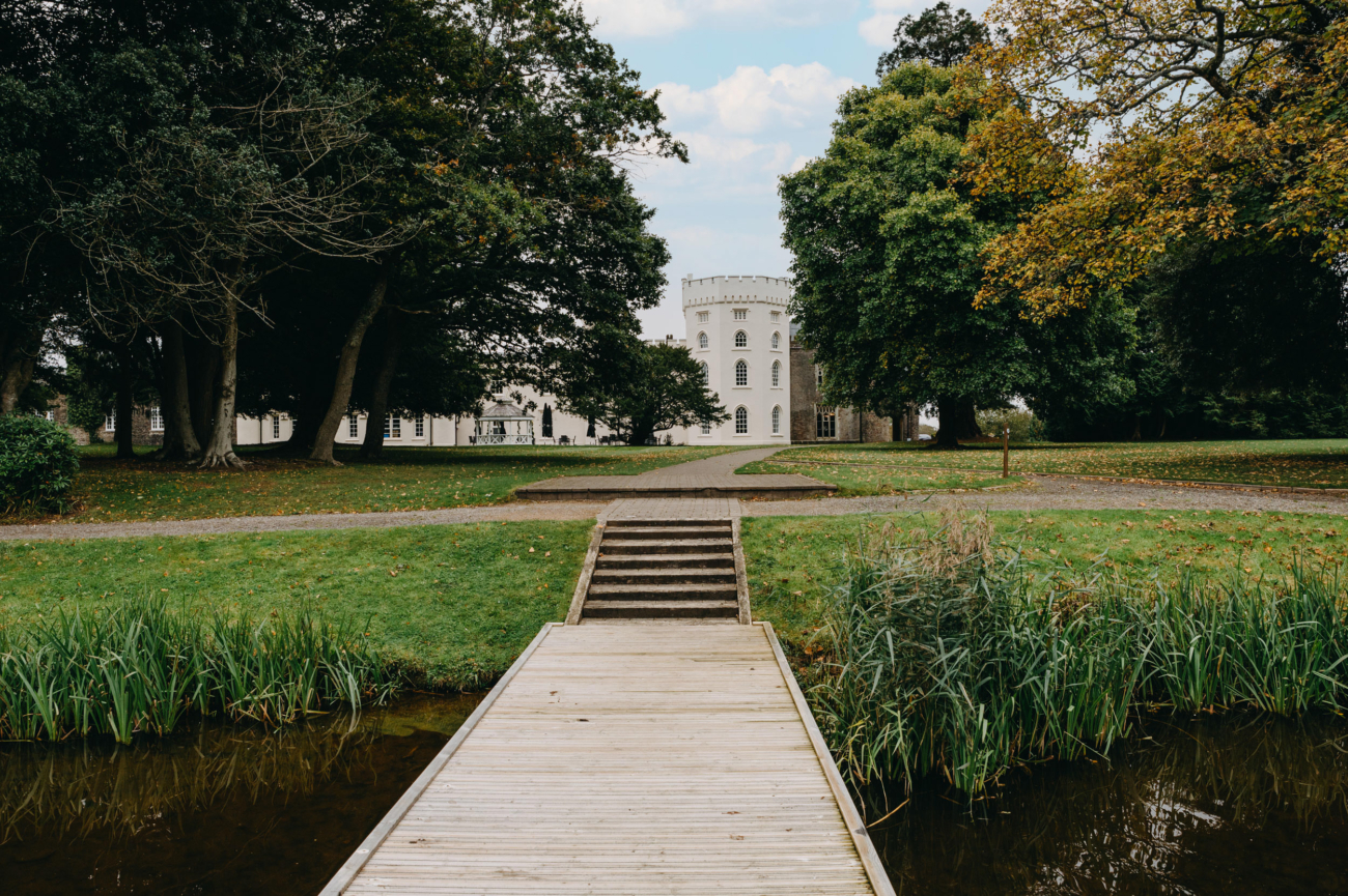 A picturesque walkway and bridge leading to the elegant Hensol Castle, a wedding venue in South Wales, surrounded by lush greenery and tranquil water.