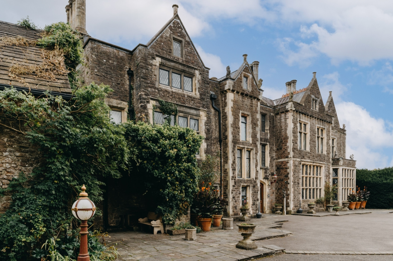 The historic facade of Miskin Manor, with ivy-covered stone walls and an inviting courtyard featuring classic potted plants and vintage lanterns.