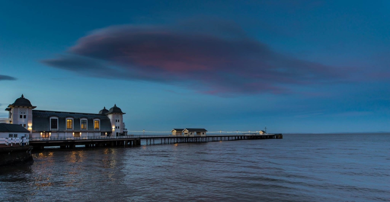 Twilight descends over Penarth Pier Pavilion, with the pavilion's warm lights glowing against a serene blue sky, creating a tranquil and romantic atmosphere by the sea.