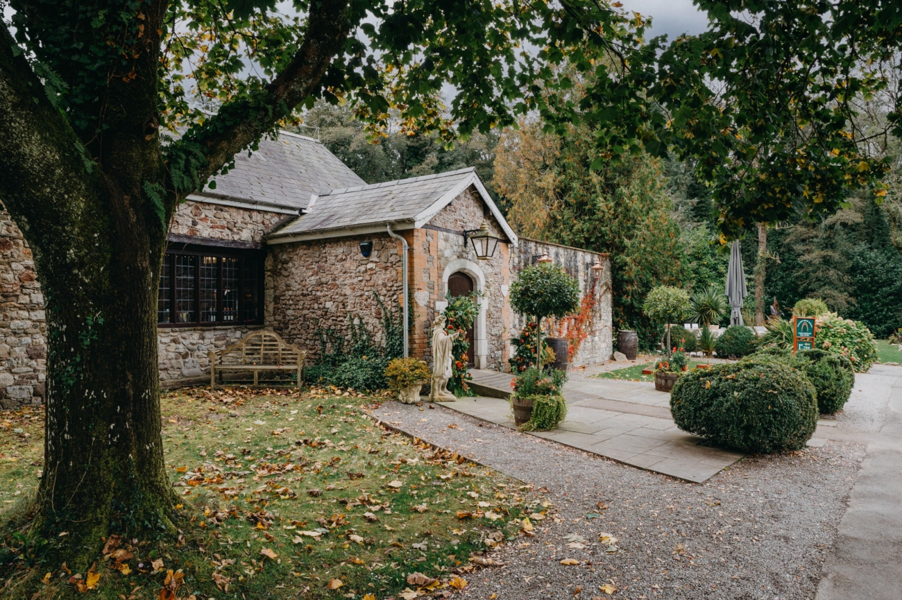 Charming entrance of Pencoed House Estate, featuring a rustic stone building surrounded by lush gardens and greenery, perfect for an intimate countryside wedding in Wales.