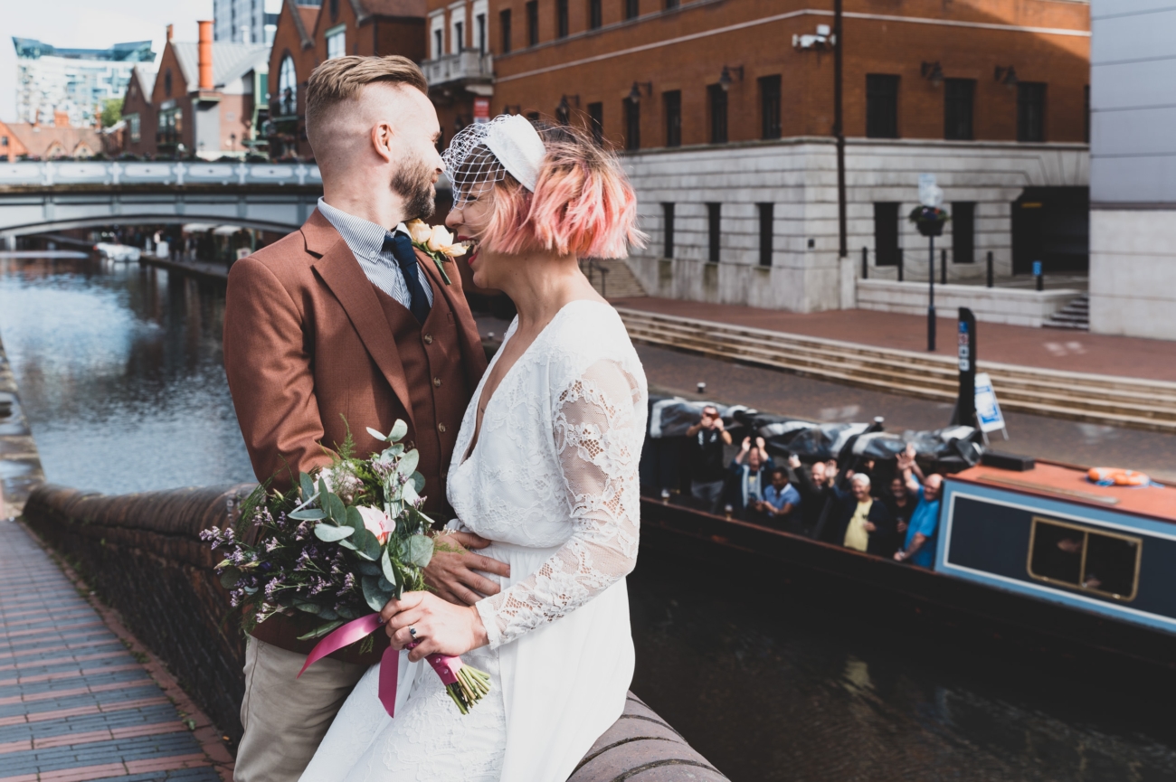 Authentic wedding photography capturing a couple by the canal in Birmingham with a joyful boat crowd in the background.