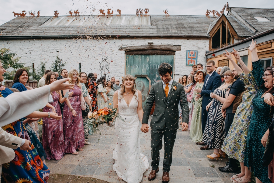 Bride and groom joyfully walk through a confetti shower, surrounded by smiling guests, symbolising the success of finding a reliable wedding photographer.