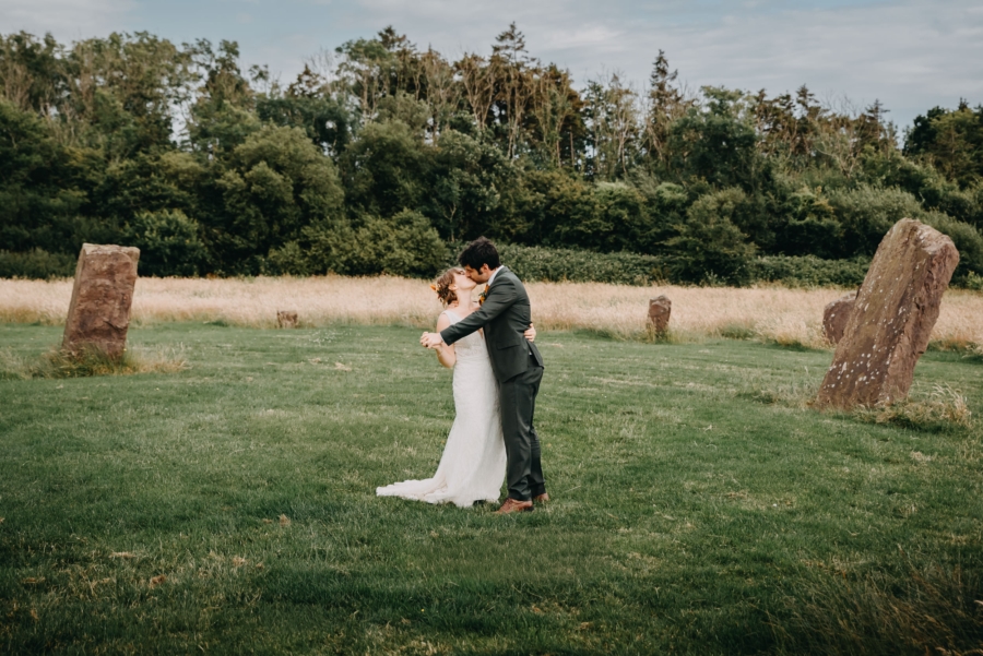 Bride and groom sharing a romantic kiss in a scenic field surrounded by ancient standing stones, beautifully captured by their wedding photographer.