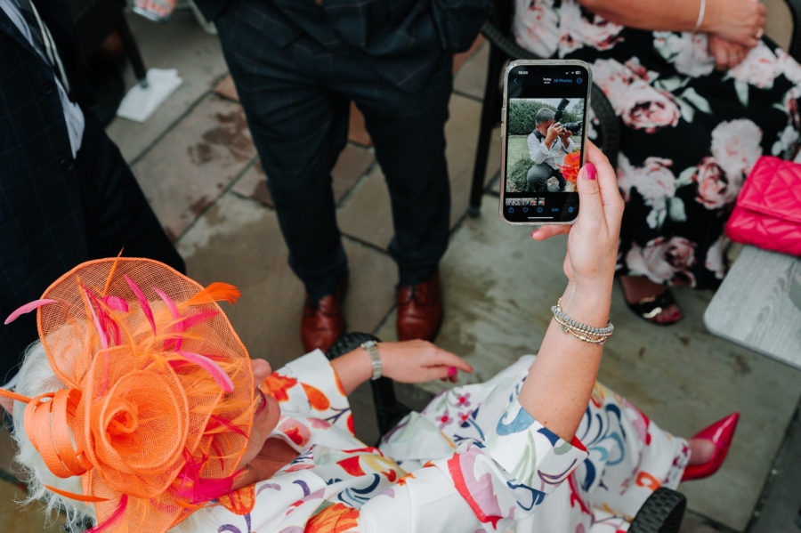 Wedding guest in a vibrant orange fascinator take a candid photo of the wedding photographer in action, capturing behind-the-scenes moments of the special day.