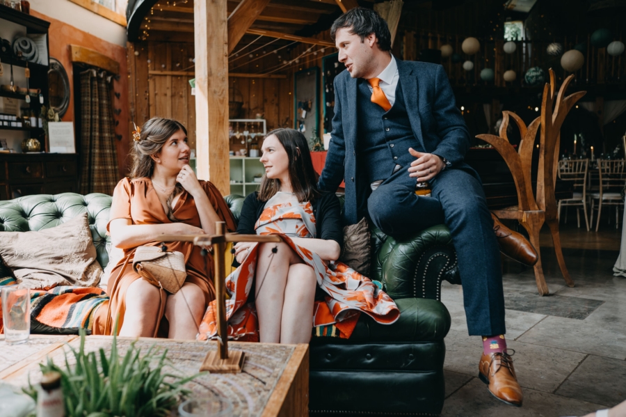 Wedding guests relaxing and chatting on a vintage green sofa in a rustic barn venue, captured by a candid wedding photographer.