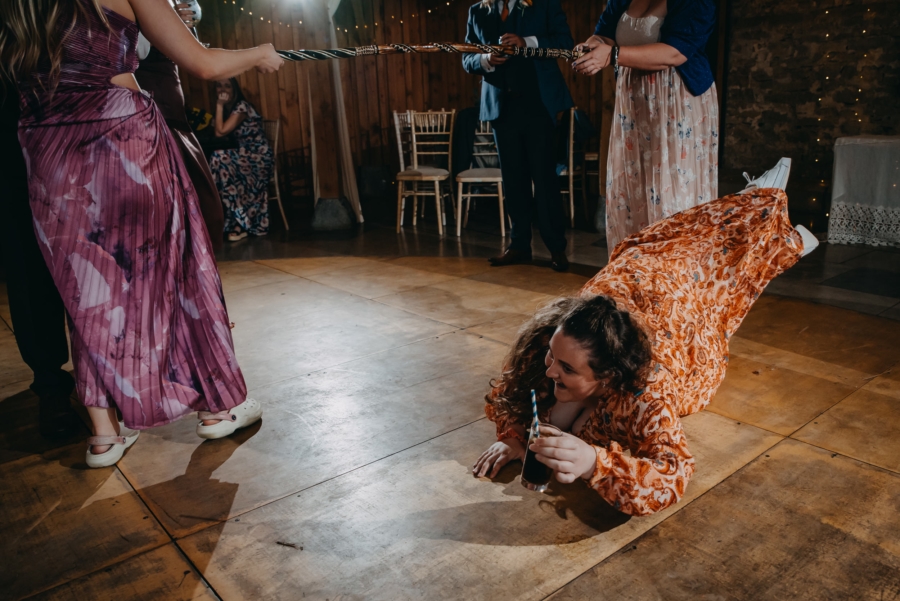 Wedding guest having fun crawling under a limbo stick with a drink in hand during a lively reception, captured by a candid wedding photographer.