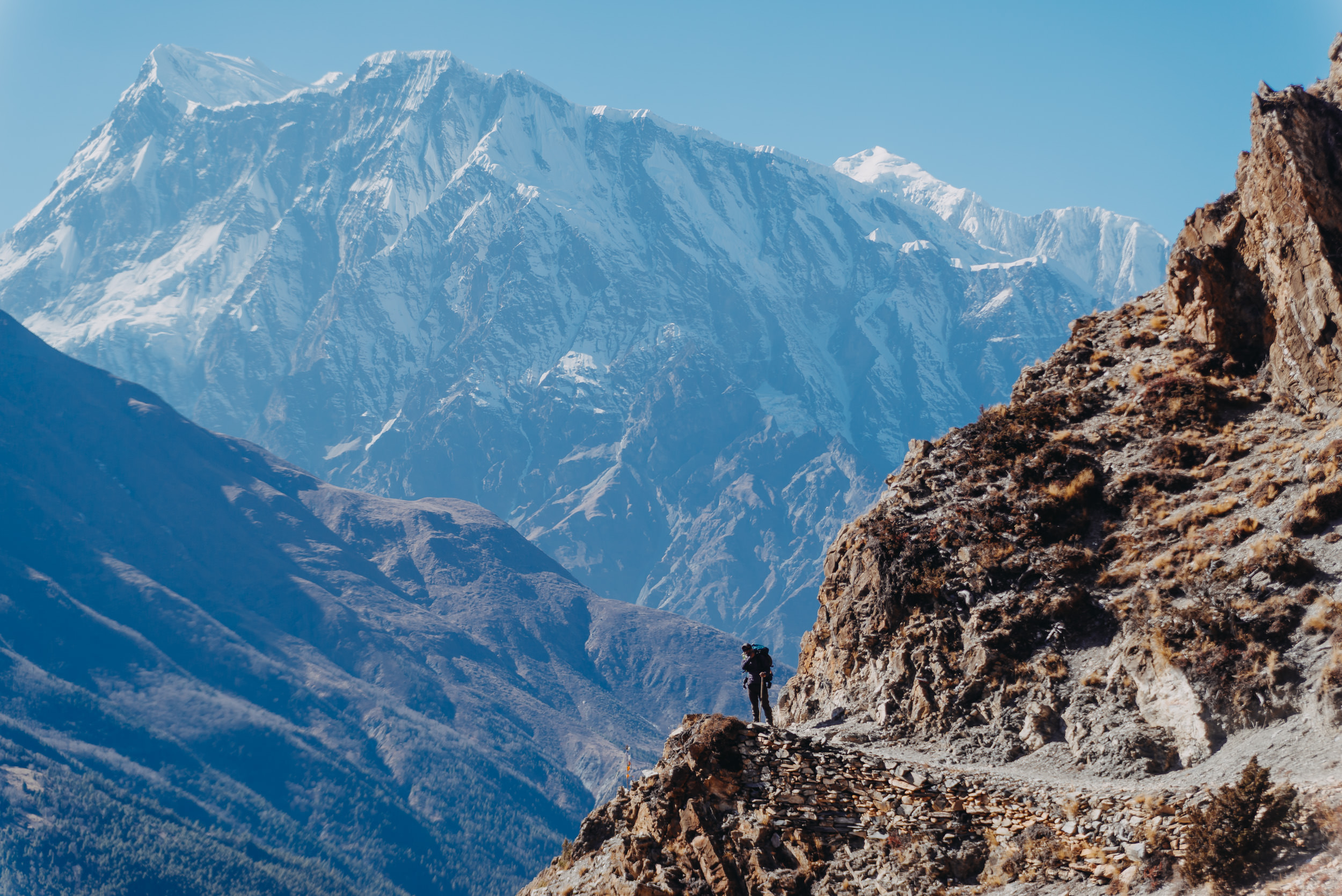 A lone trekker standing on a narrow mountain trail along a rocky cliff, with a majestic view of snow-capped Annapurna II mountains in the background.