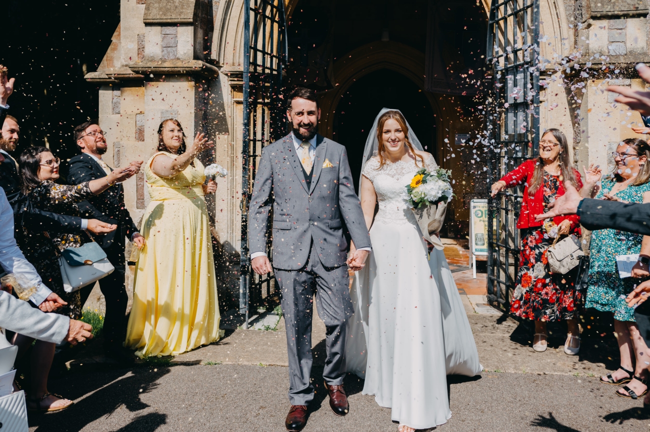 Newlyweds walking hand in hand through confetti outside All Saints Church, Babbacombe, Torquay.