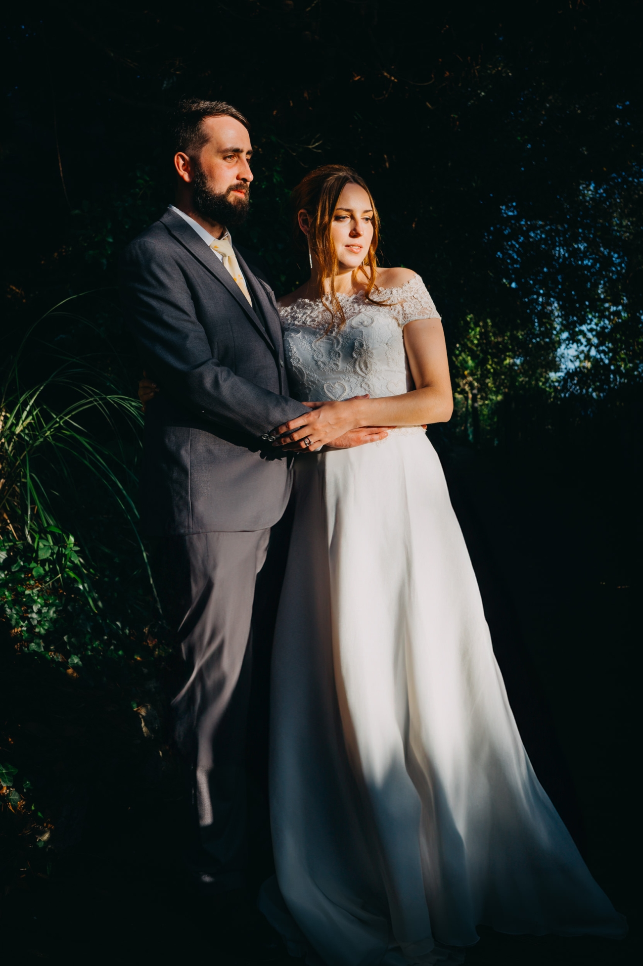 Bride and groom standing together in golden sunlight, holding hands with a natural green backdrop.