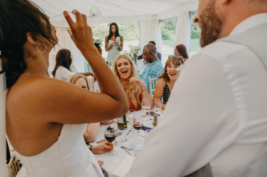 Bride drinking wine while wedding guests burst into laughter – candid reception moment.