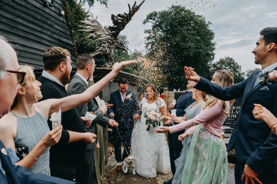 Bride and groom walking through confetti with their dog at an alternative wedding venue featuring a striking wooden sculpture.