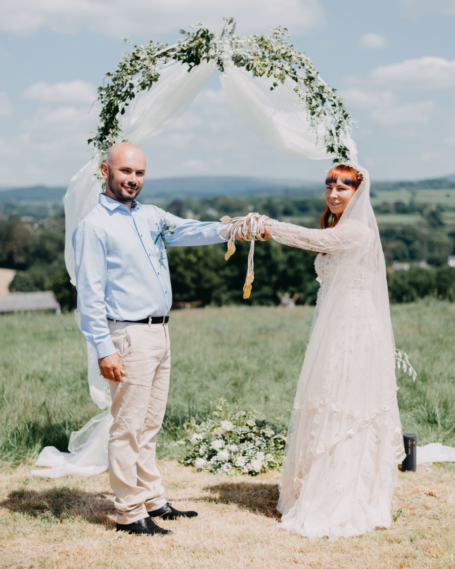 Bride and groom holding hands with cords during symbolic wedding ceremony outdoors