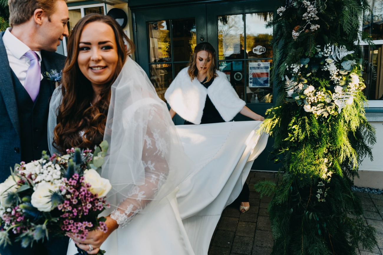 Bride and groom outside Myddfai Community Hall with bridesmaid adjusting the dress – moment captured by a documentary wedding photographer