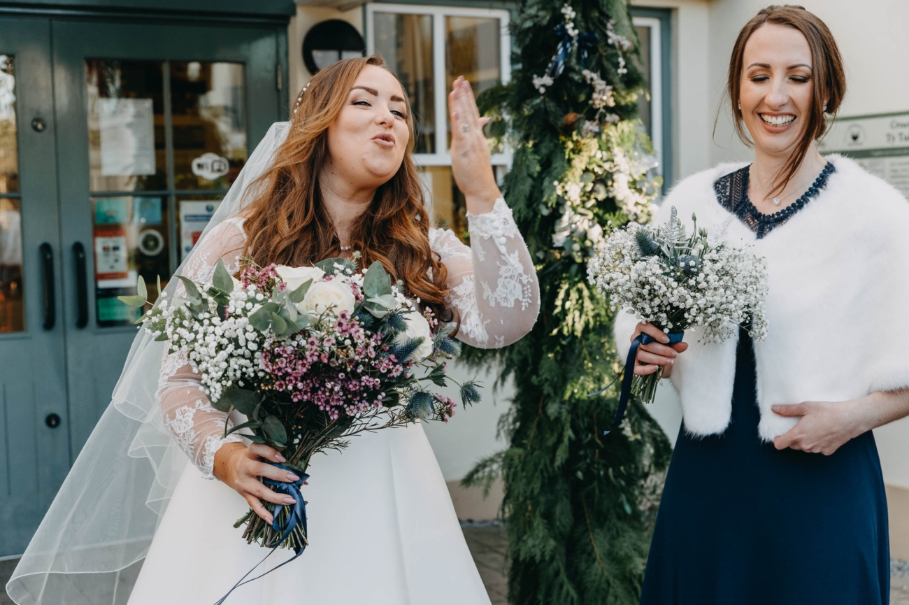 Bride blowing a kiss outside Myddfai Community Hall – candid moment after the ceremony with bridesmaid smiling
