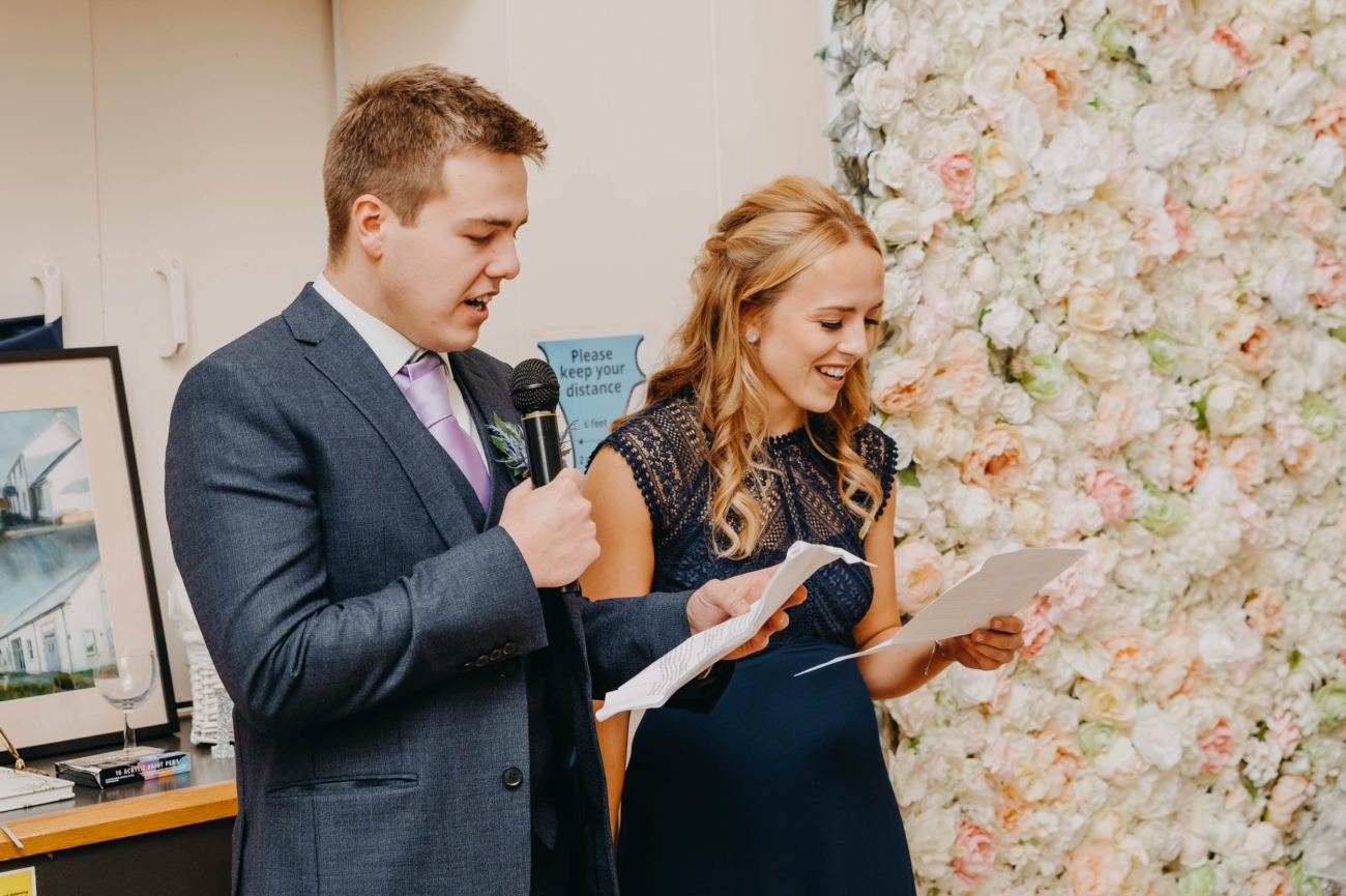 Two wedding guests giving a joint speech during the reception at Myddfai Community Hall – smiling and candid moment